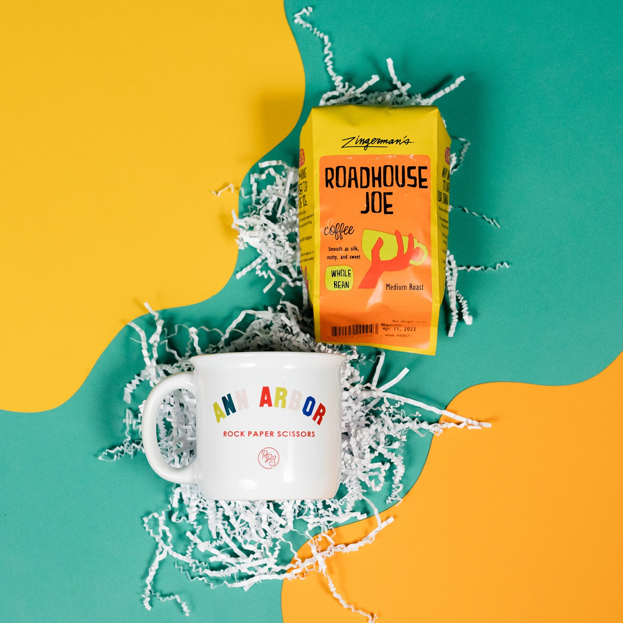 On a sunny mustard yellow and cool aqua backdrop lays a mug and a bag of coffee. The mug is a large white mug with colorful collegiate font that says "Ann Arbor" with "ROCK PAPER SCISSORS" below it. The bright yellow blue bag of coffee lays next to the mug. The coffee is Zingerman's brand Roadhouse Joe Blend. 'Smooth as silk and served daily at Zingerman's Roadhouse' Ground coffee. 12 oz (350 g). White crinkle is scattered around.