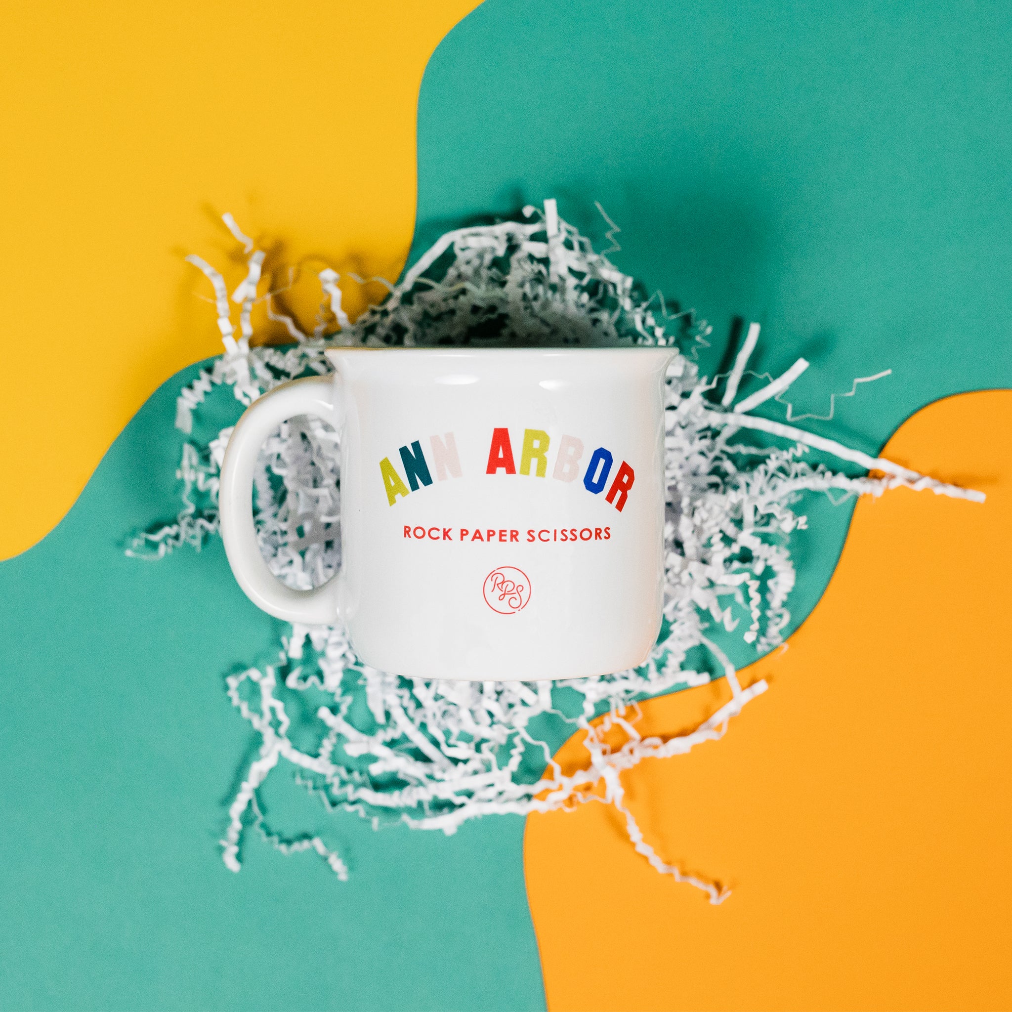 On a sunny mustard yellow and cool aqua backdrop lays a large white mug with colorful collegiate font that says "Ann Arbor" with "ROCK PAPER SCISSORS" below it. White crinkle surrounds the mug.