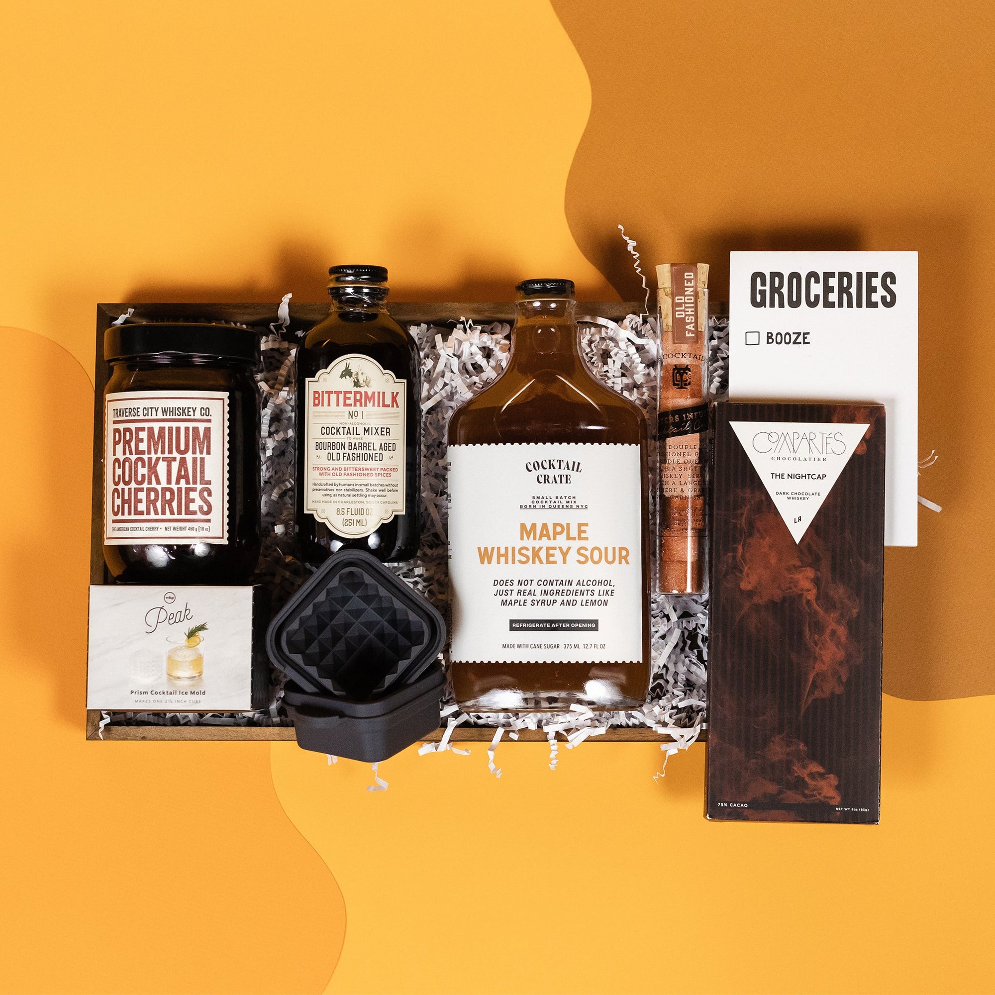On a retro mustard yellow and ochre background sits a wooden tray of whiskey-themed gifts with white paper krinkle. The gifts include a small bottle of Bittermilk Bourbon Barrel Aged Old Fashioned cocktail mix, Cocktail Crate Maple Whiskey Sour mixer, a Peak Prism Cockail Ice Mold, Traverse City Whiskey Co. Premium Cocktail Cherries, YES Cocktail Co. Old Fashioned Sugar Cubes, a "GROCERIES" notepad with one checkbox for Booze, and a premium Compartes 'The Nightcap' Dark Chocolate Whiskey Bar. 