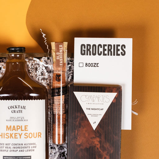 On a retro mustard yellow and ochre background sits a wooden tray of whiskey-themed gifts with white paper krinkle. This photo is a close-up of the white "GROCERIES" notepad with one checkbox for Booze, Cocktail Crate Maple Whiskey Sour mixer, and premium Compartes 'The Nightcap' Dark Chocolate Whiskey Bar. 