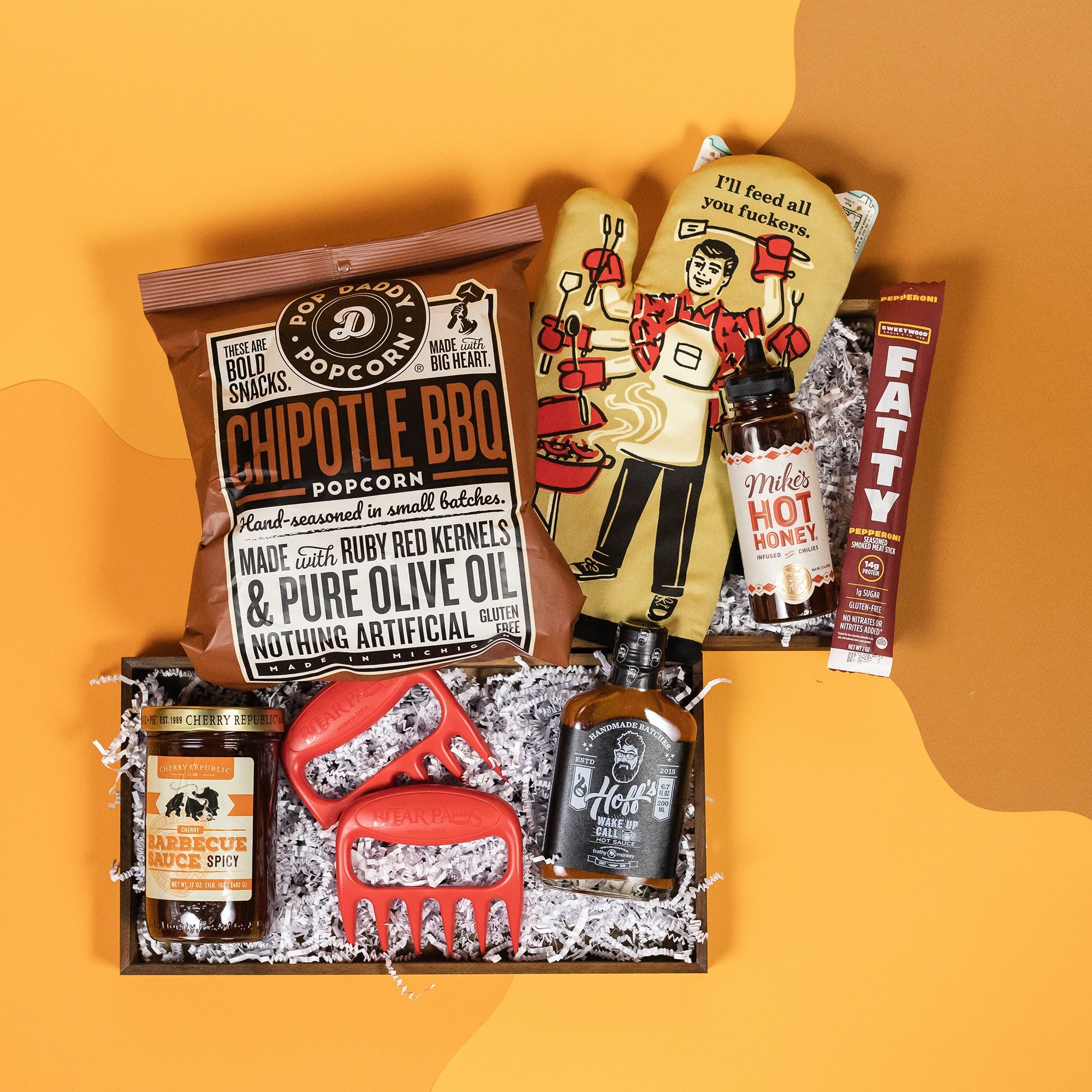 On a retro mustard yellow and ochre background sits a wooden tray of BBQ-themed gifts with white paper krinkle. The products are tied together with a cohesive red-brown-black color story. The products include a red set of Bearpaw meat claws for shredding, an oven mitt with a retro 50's dad illustration that says "I'll Feed All You Fuckers," Mike's Hot Honey, a Fatty Pepperoni stick, Hoff's Wake Up Call Hot Sauce, Cherry Republic Spicy BBQ Sauce and a bag of Pop Daddy Chipotle BBQ Popcorn.