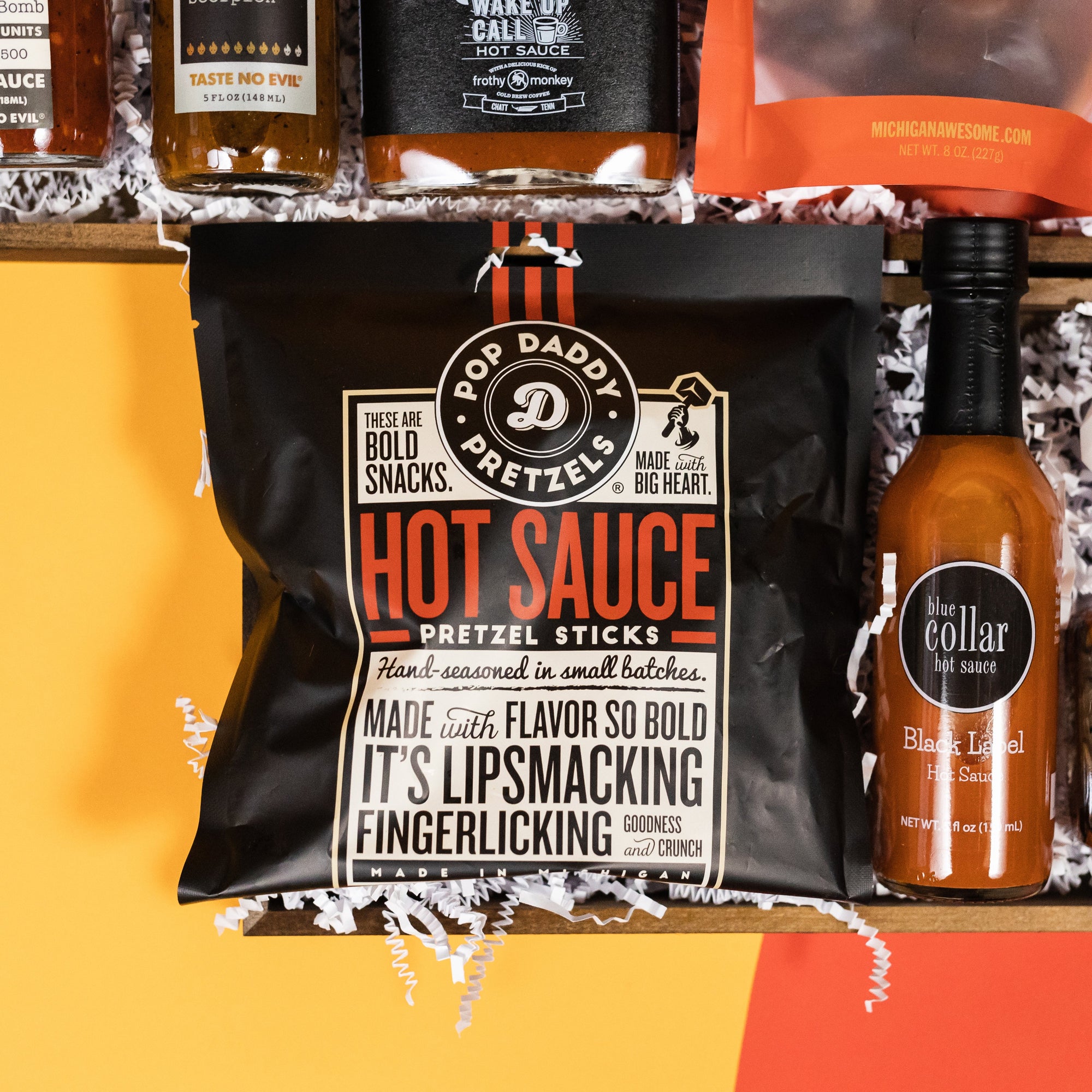 On a bright yellow backgorund with orange and red cutouts sit two offset wooden trays filled to the brim with locally-made and small batch hot sauce themed porducts and white paper krinkle. This photo is a close-up of Blue Collar Black Label Hot Sauce and a bag of Pop Daddy Hot Sauce Pretzel Sticks.