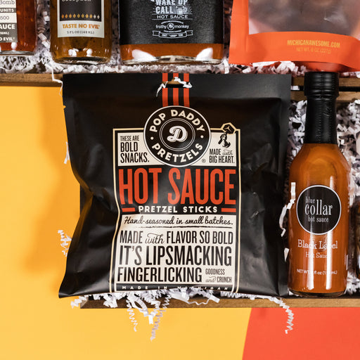 On a bright yellow backgorund with orange and red cutouts sit two offset wooden trays filled to the brim with locally-made and small batch hot sauce themed porducts and white paper krinkle. This photo is a close-up of Blue Collar Black Label Hot Sauce and a bag of Pop Daddy Hot Sauce Pretzel Sticks.