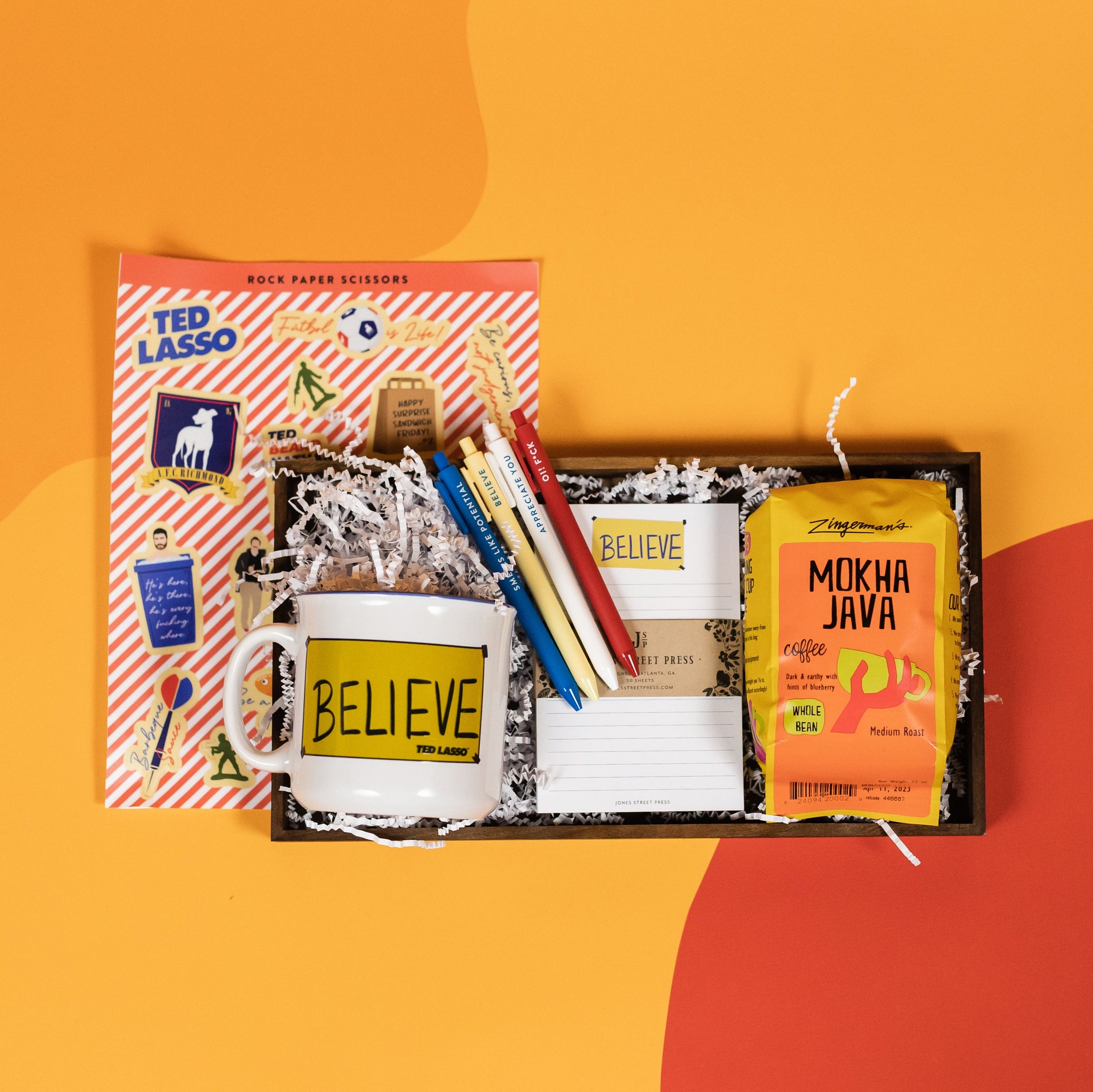 On a bright yellow background with orange and red cutouts sits a wooden tray with an assortment of Ted Lasso themed gifts and white krinkle. The products include a white mug with yellow "BELIEVE" banner illustrated on the side, an RPS Exclusive Ted Lasso sticker sheet, a set of four retractable gel pens with Ted Lasso sayings, a white lined notepad with the illustrated "BELIEVE" banner at the top and a bag of Zingerman's Mokha Java Medium Roast Whole Bean Coffee.