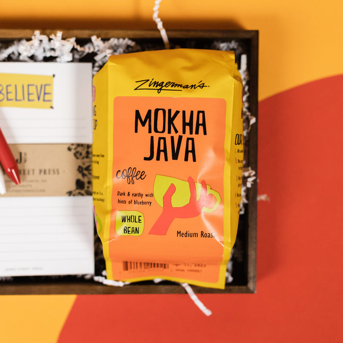 On a bright yellow backgorund with orange and red cutouts sits a wooden tray with an assortment of Ted Lasso themed gifts and white krinkle. This photo is a close-up of a bag of Zingerman's Mokha Java Medium Roast Whole Bean Coffee.