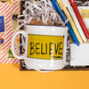 On a bright yellow background sits a wooden tray with an assortment of Ted Lasso themed gifts and white krinkle. This photo is a close-up of a white mug with yellow "BELIEVE" banner illustrated on the side.