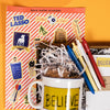 On a bright yellow background with orange and red cutouts sits a wooden tray with an assortment of Ted Lasso themed gifts and white krinkle. This photo is a close-up of the RPS Exclusive Ted Lasso sticker sheet and set of four retractable gel pens with Ted Lasso sayings.