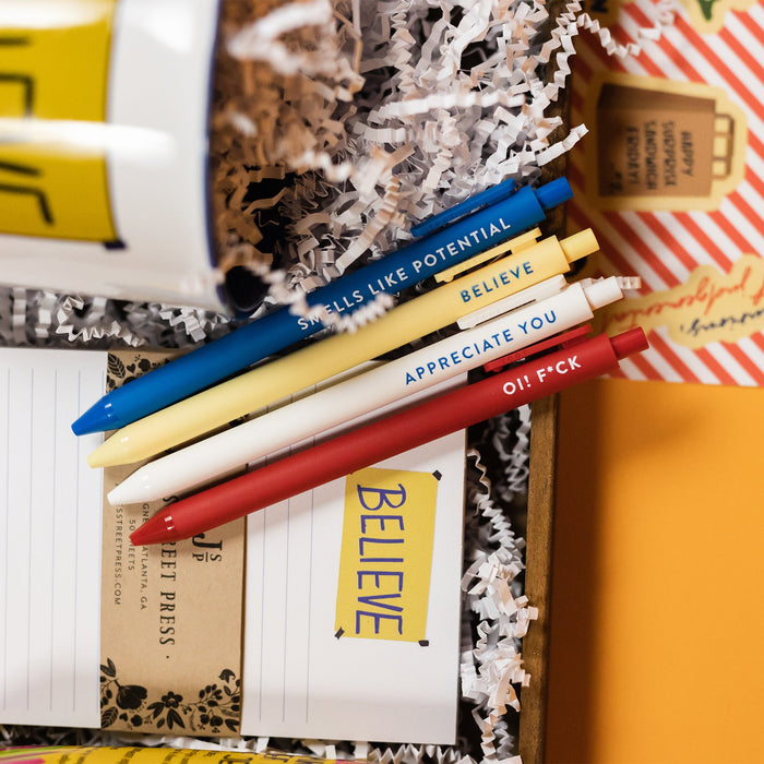 On a bright yellow background sits a wooden tray with an assortment of Ted Lasso themed gifts and white krinkle. This photo is a close-up of the set of four retractable gel pens with Ted Lasso sayings. It says "SMELLS LIKE POTENTIAL," "BELIEVE," "APPRECIATE YOU," and "OI! F*CK." They are in blue, light yellow, white, and red.