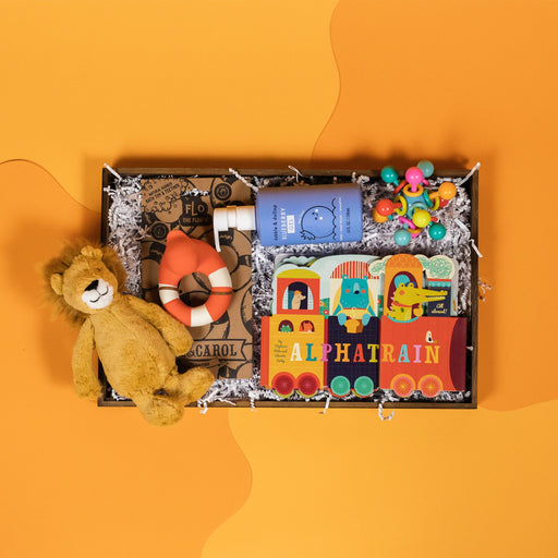 On a funky yellow and yellow-orange background sits a wooden tray filled to the brim with rainbow baby toys. The gifts include a super-soft lion plushie, a red teether bath duckie floatie, a blue bottle of Blueberry Gel tear-free bubbles, Alphatrain board book by Stephanie Miles and Christin Farley and a Manhattan Toy Atom Teether.