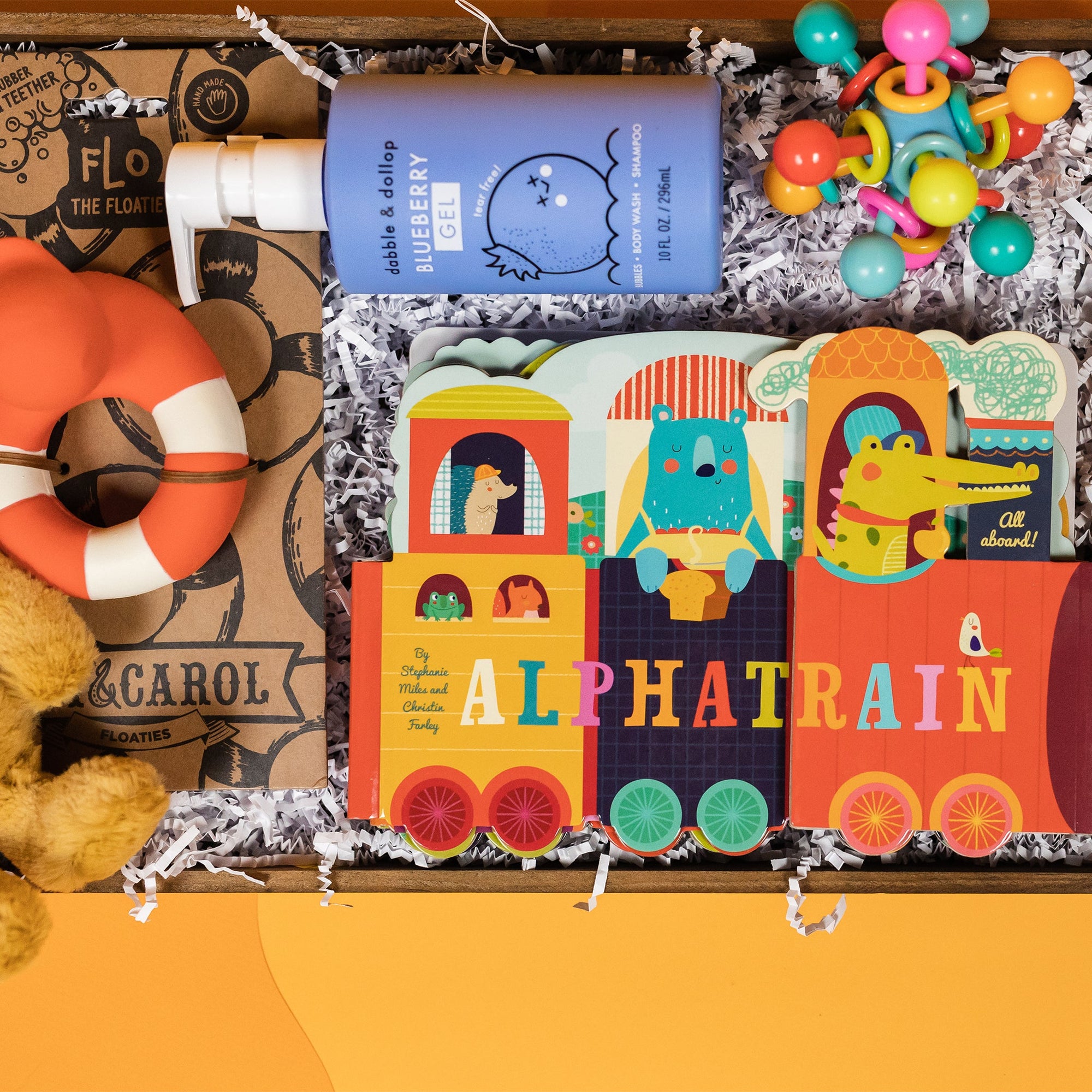 On a funky yellow and yellow-orange background sits a wooden tray filled to the brim with rainbow baby toys. This photo is a close-up of a blue bottle of Blueberry Gel tear-free bubbles, Alphatrain board book by Stephanie Miles and Christin Farley and a Manhattan Toy Atom Teether..