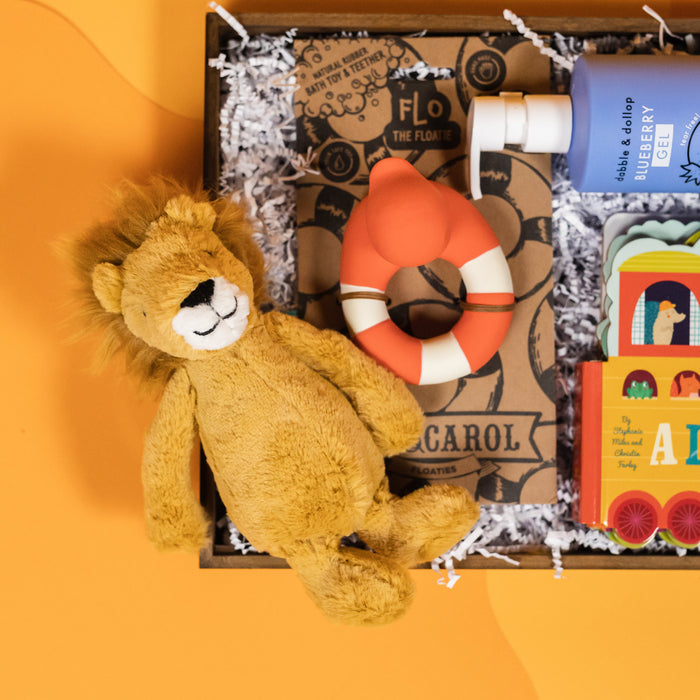On a funky yellow and yellow-orange background sits a wooden tray filled to the brim with rainbow baby toys. This photo is a close-up of a super-soft lion plushie and a red teether bath duckie floatie.