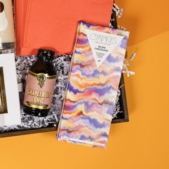On a funky light orange and sunny mustard background sits a wooden tray filled to the brim with brunch accoutrements. This photo is a close-up of  a small bottle of Portland Syrups Grapefruit Tonic drink mixer and a gorgeous Compartes Tie Dye White Chocolate Bar.