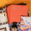 On a funky light orange and sunny mustard background sits a wooden tray filled to the brim with brunch accoutrements. This photo is a close-up of  a stack of coral beverage napkins that say "STARTED FROM THE BOTTOMLESS MIMOSAS NOW WE HERE" in silver.