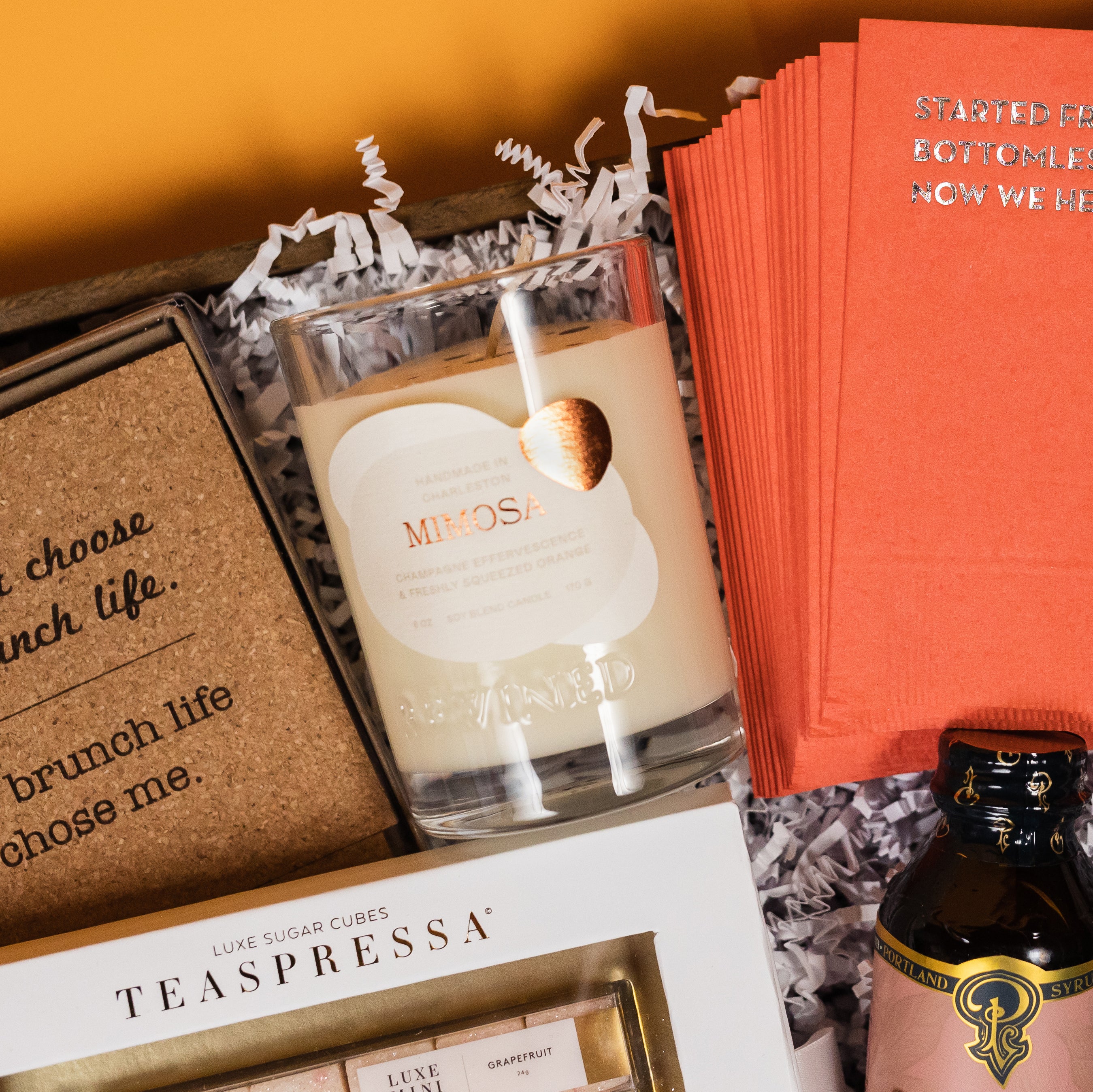 On a funky light orange background sits a wooden tray filled to the brim with brunch accoutrements. This photo is a close-up of  a premium soy blend Mimosa candle that smells of 'champagne effervescence and freshley squeezed orange'.