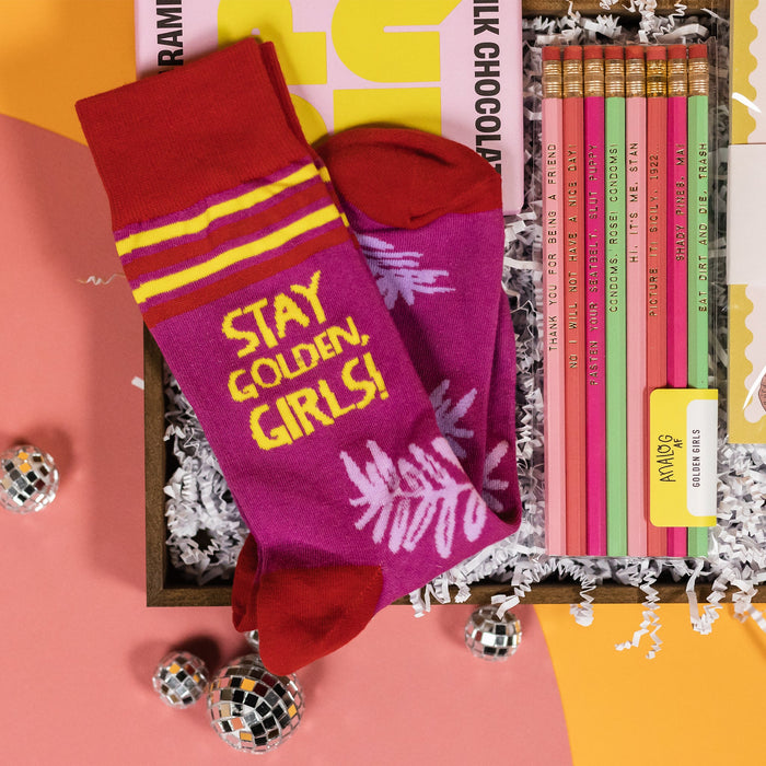 On a coral and sunny mustard wavy background with small silver disco balls sits a wood box full of Golden Girls inspired gifts. It is a close-up of a pair of purple, dark red, and yellow socks. It says "STAY GOLDEN, GIRLS!" in yellow, all caps handwritten lettering.  There is also a clear package with a set of 8 pencils in baby pink, coral, hot pink, and mint. They have various sayings on them from the hit show. Under the socks is a box of UP UP Milk Chocoloate. The package is in baby pink and yellow.