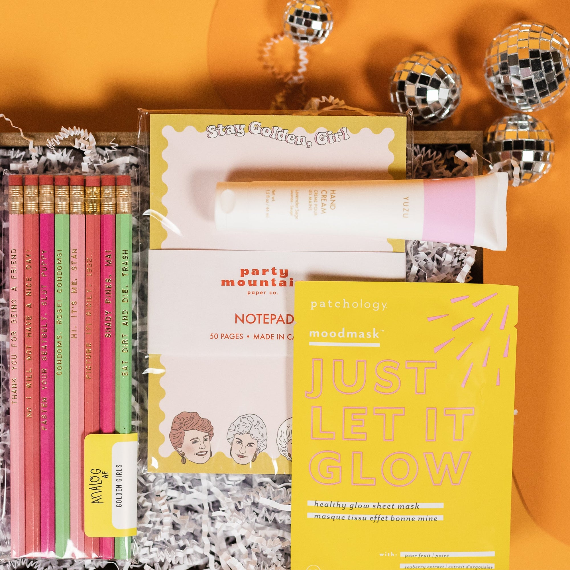 On a sunny mustard and orange wavy background with small silver disco balls sits a wood box full of Golden Girls inspired gifts. It is close-up a clear package with a set of 8 pencils in baby pink, coral, hot pink, and mint. They have various sayings on them from the hit show. Next to it is a "Stay Golden, Girl" white notepad, white tube of 'Yuzu' Hand Cream, and a yellow package of "JUST LET IT GLOW" healthy glow sheet mask with pear fruit.