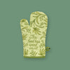 On a mossy green background sits an oven mitt. This celery green mitt is covered with illustrations of marijuana leaves and leaves in mid green. It says "The food has weed in it" in handwritten, script lettering in mid green.