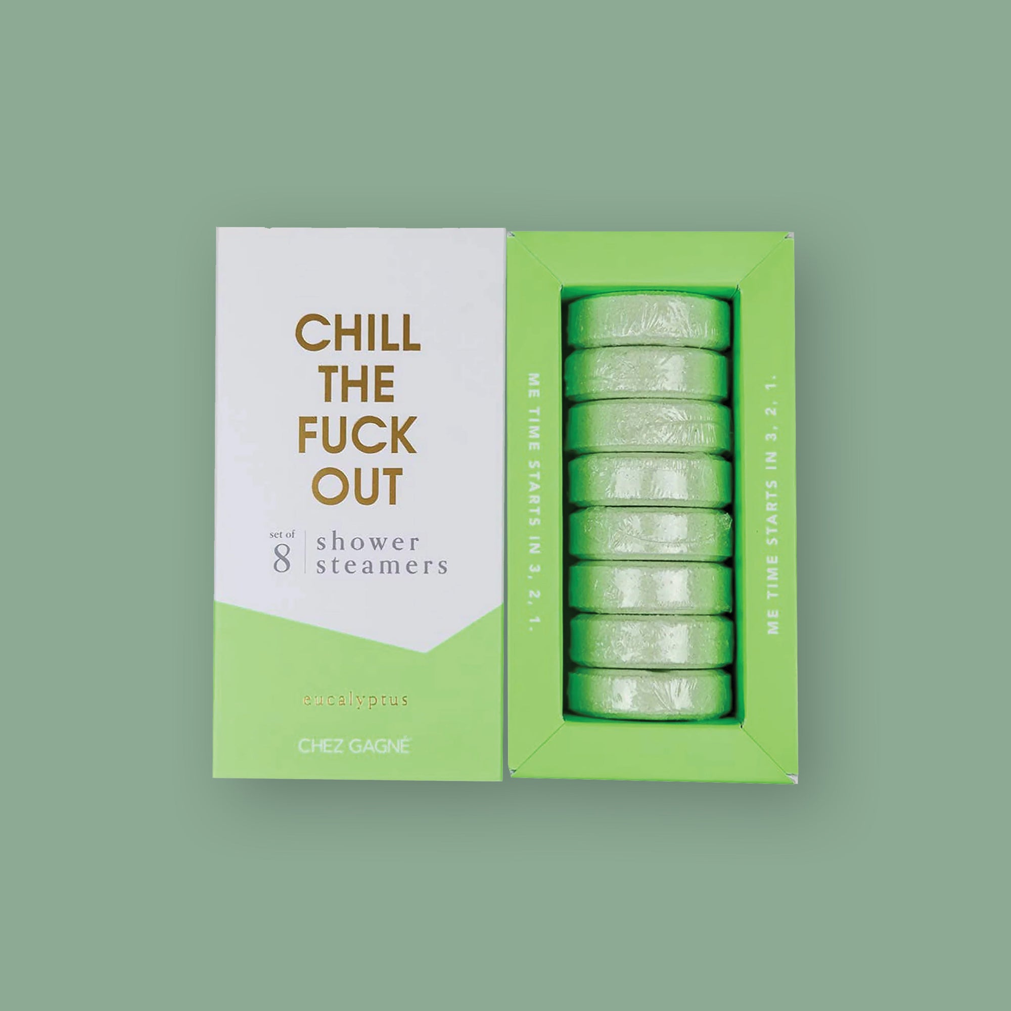 On a mossy green background sits two boxes. This picture is a close-up of a white and pistachio green package that says "CHILL THE FUCK OUT" in gold foil, all caps block lettering. Under it says "set of 8" and " shower steamers" in grey, lowercase serif font. At the bottom it says "eucalyptus" in gold foil, lower case serif font. To the right of it is a pistachio green box that is out of focus and has pistachio green shower steamers in it.