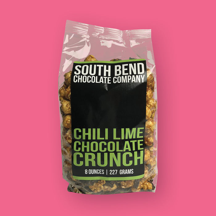 On a hot pink background sits a package. This clear package has a black label on it. It says "SOUTH BEND CHOCOLATE COMPANY" in white, all caps block font. On the bottom it says "CHILI LIME CHOCOLATE CRUNCH" in lime green, all caps block font. It is filled with popcorn. 8 ounces | 227 grams