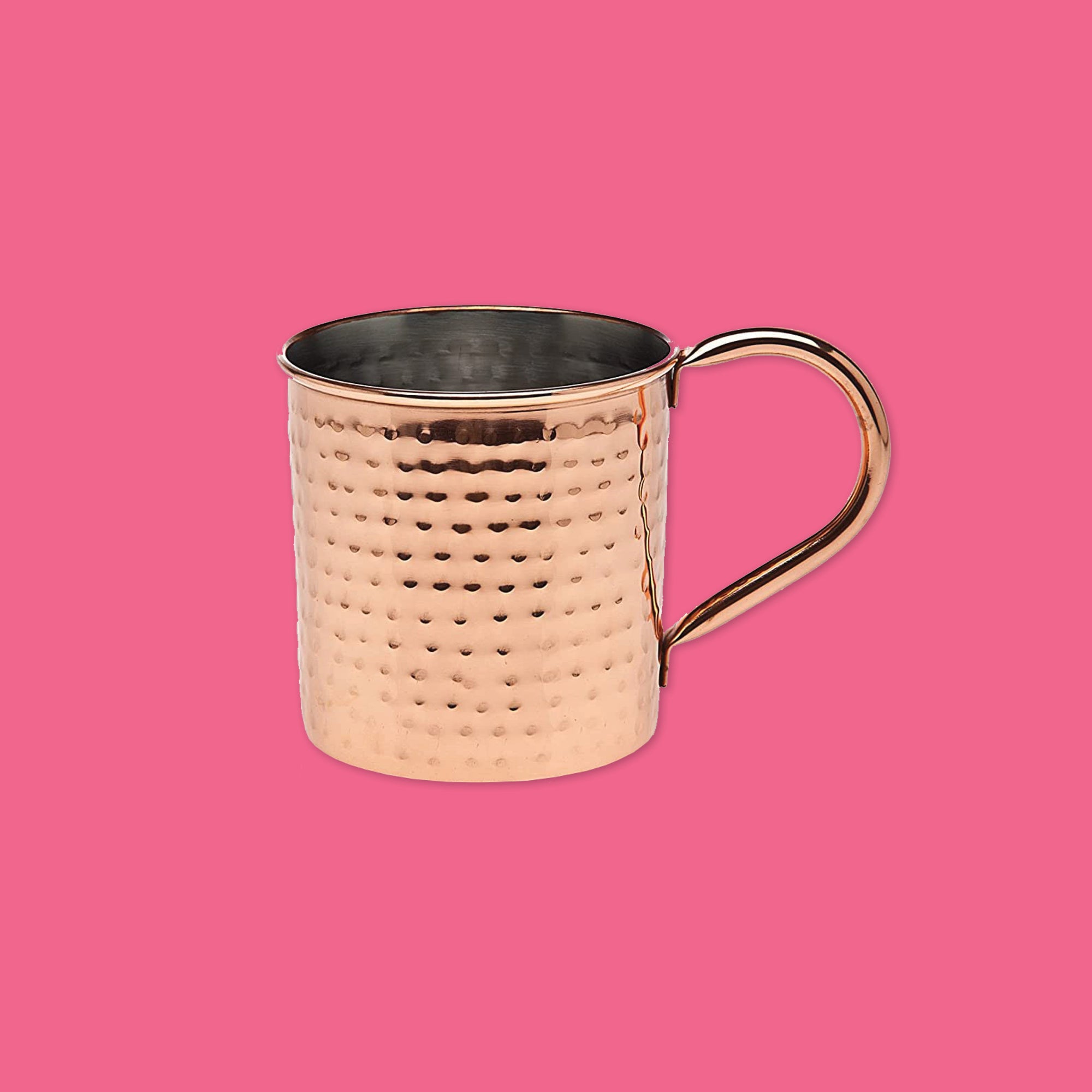 On a hot pink background sit a hammered, copper mug. 