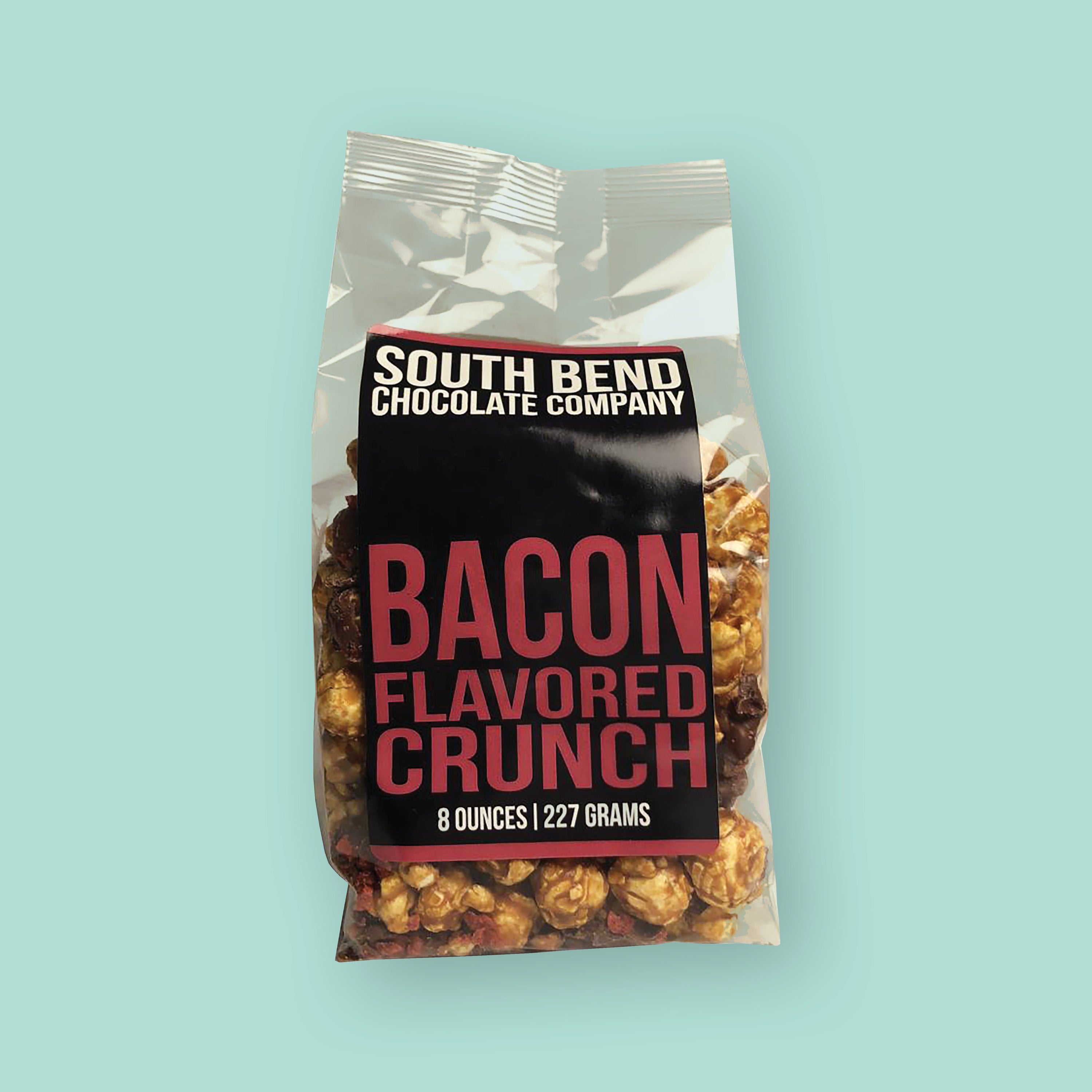 On a mint green background sits a package. This clear package is filled with caramel popcorn and has a black label. It says "SOUTH BEND CHOCOLATE COMPANY" in white, all caps block font. It says "BACON FLAVORED CRUNCH" in a maroon, all caps block font. 8 ounces | 227 grams