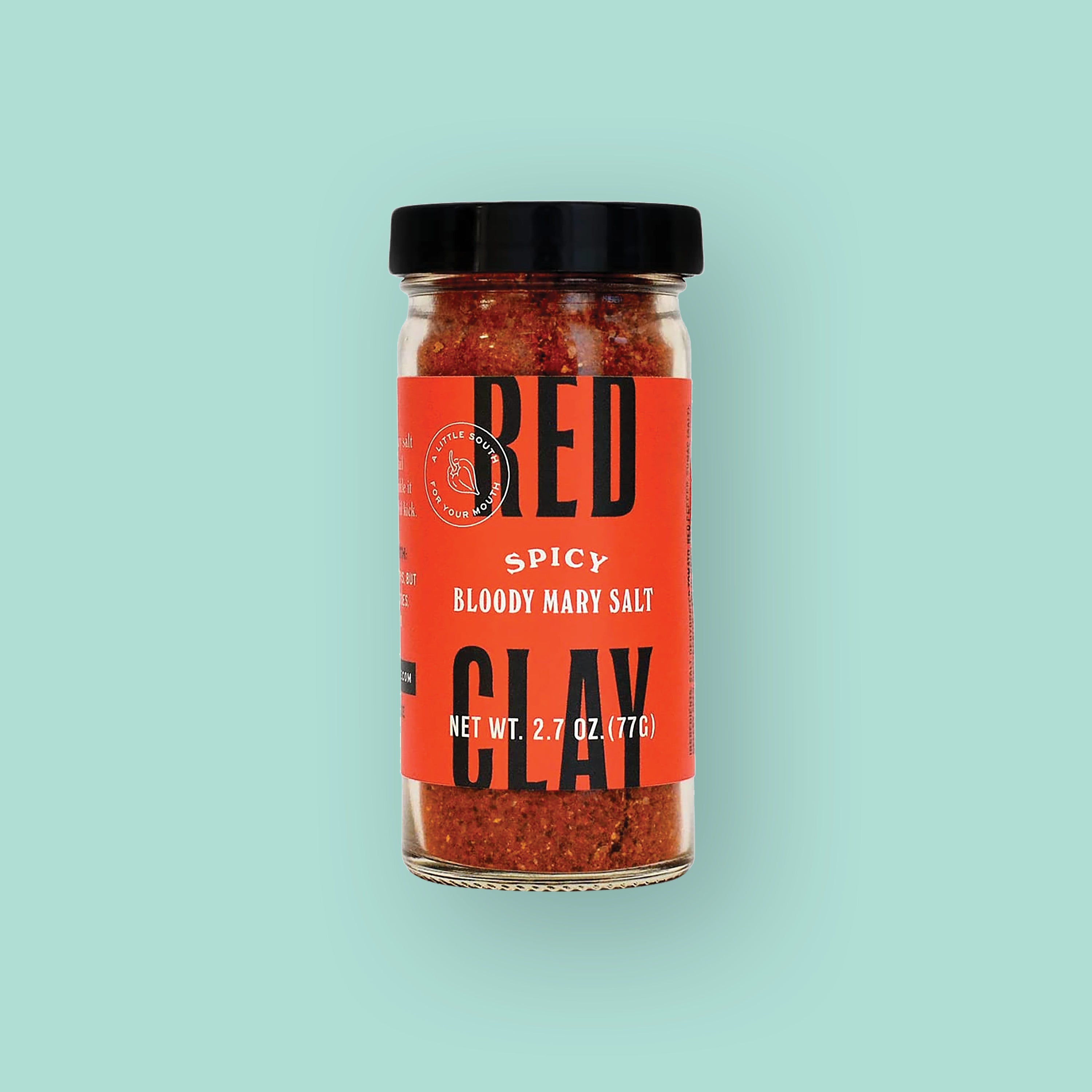 On a mint green background sits a bottle. This glass bottle has a black lid and red label. It says "RED CLAY" in black, all caps bold font and "SPICY BLOODY MARY SALT" in white, all caps serif font. It is filled with reddish seasoning. NET WT. 2.7 OZ. (77G)