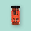 On a mint green background sits a bottle. This glass bottle has a black lid and red label. It says "RED CLAY" in black, all caps bold font and "SPICY BLOODY MARY SALT" in white, all caps serif font. It is filled with reddish seasoning. NET WT. 2.7 OZ. (77G)