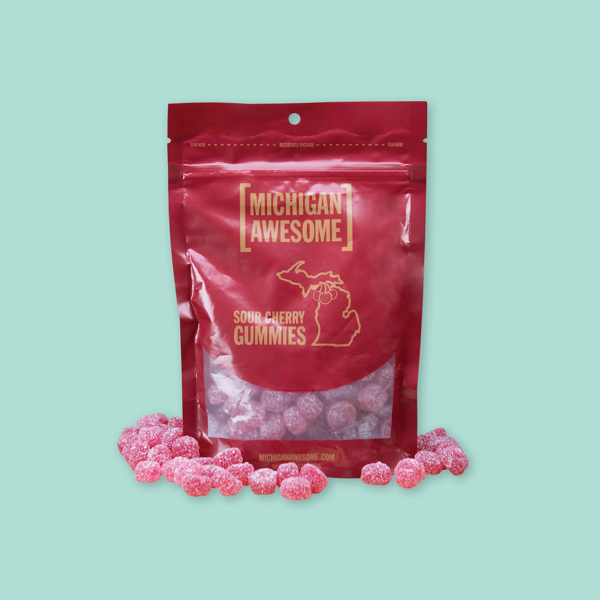 On a mint green background sits a red package that is see-through at the bottom. It is filled with sugar sour cherry gummies. It says "MICHIGAN AWESOME," and "SOUR CHERRY GUMMIES" in goldish-yellow, all caps block font. It has an outlined illustration of the state of Michigan-UP with cherries. There are sugar sour cherry gummies sitting around the bag.