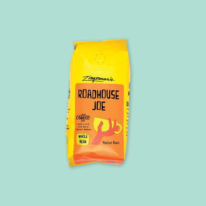 On a mint green background sits a coffee package. This yellow package has an orange label on the front. It says "Zingerman's" at the top in black, handwritten lettering. It also says "ROADHOUSE JOE" in black, handwritten lettering with an illustration of a hand holding a yellow mug. It also says "coffee Smooth as silk & served daily at Zingerman's Roadhouse" in black, handwritten lettering. Whole bean, medium roast 12 oz