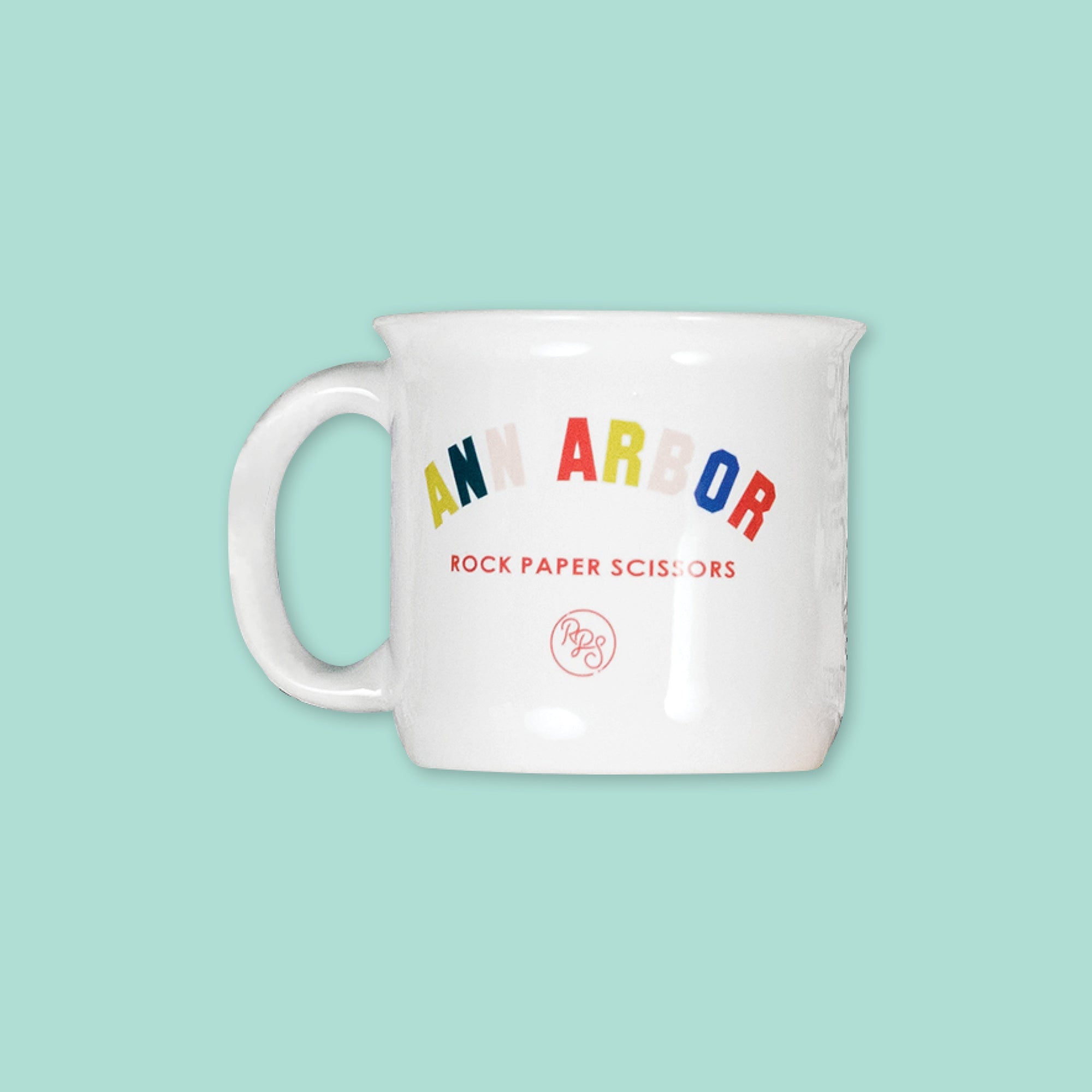 On a mint green background sits a mug. The white mug has a color collegiate font that says "ANN ARBOR" and under it says "ROCK PAPER SCISSORS" in a red, all caps block font. Under that is a circular, red "RPS" logo in handwritten script lettering.