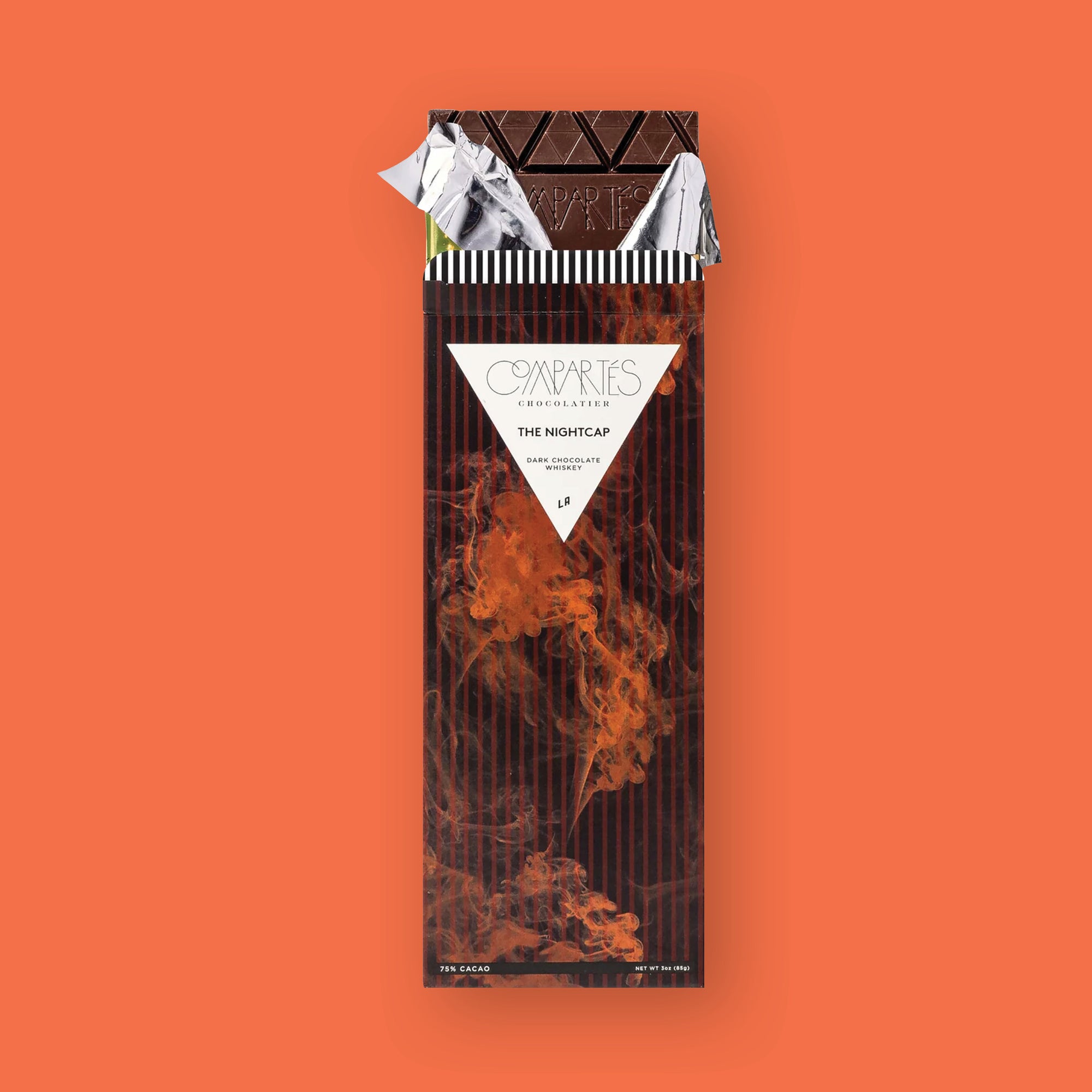 On an orangey-red background sits an opened package. This brown striped, smokey package has a chocolate bar sticking out of the top with gold and silver foil that is opened. On the front of the package is a white triangle and it says "COMPARTES CHOCOLATIER," "THE NIGHTCAP," "DARK CHOCOLATE WHISKEY," and "LA" all in black font. 75% CACAO NET WT 3OZ (85G)