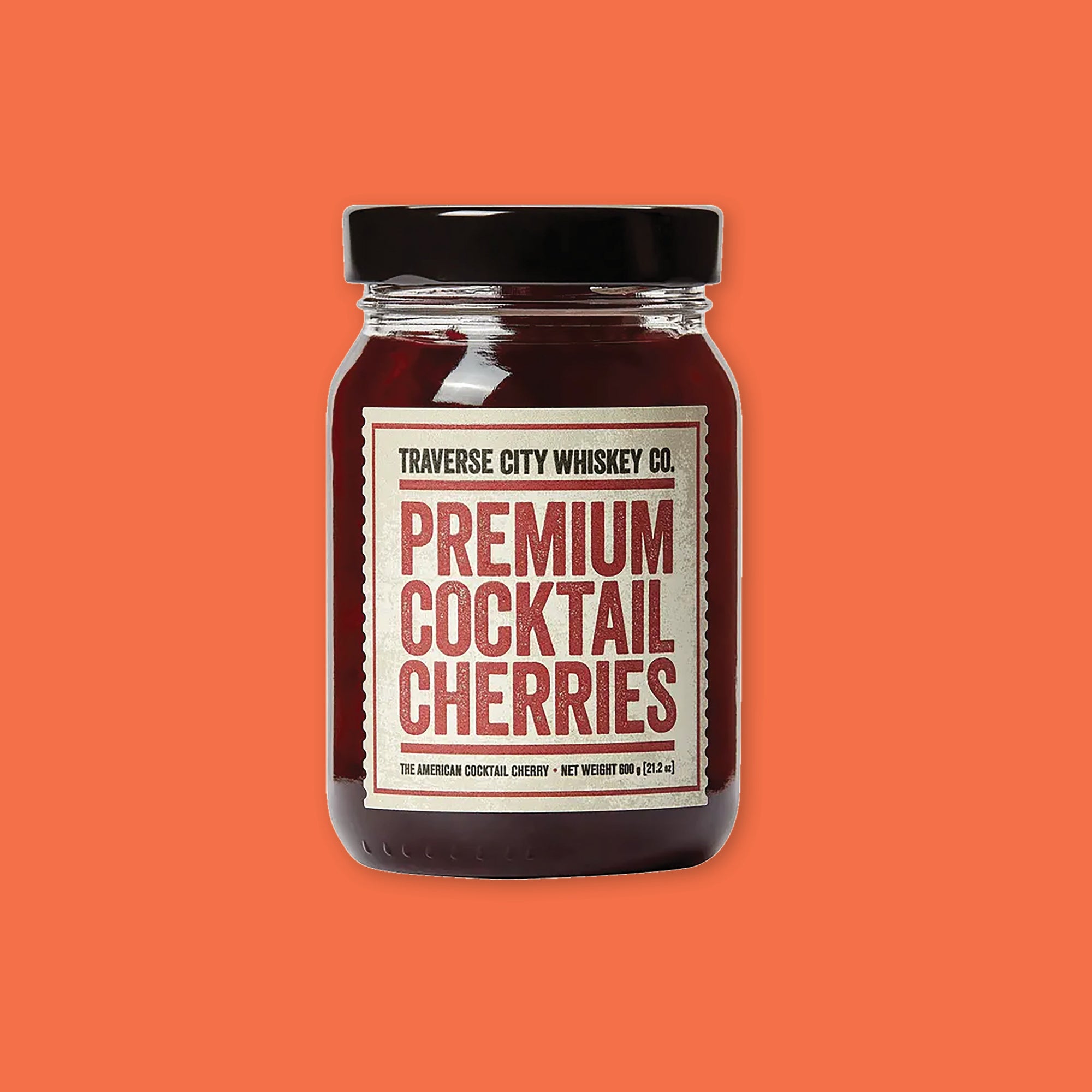 On an orangey-red background sits a jar. The glass jar has a black lid and and a vintage creamy label that says "TRAVERSE CITY WHISKEY CO." in black, all caps block font. Under it is a red line and it says "PREMIUM COCKTAIL CHERRIES" in a red, all caps block font. There is another red line under that and then it says "THE AMERICAN COCKTAIL CHERRY • NET WEIGHT 600 g (21.2 oz)." There is a red outline square around the copy.