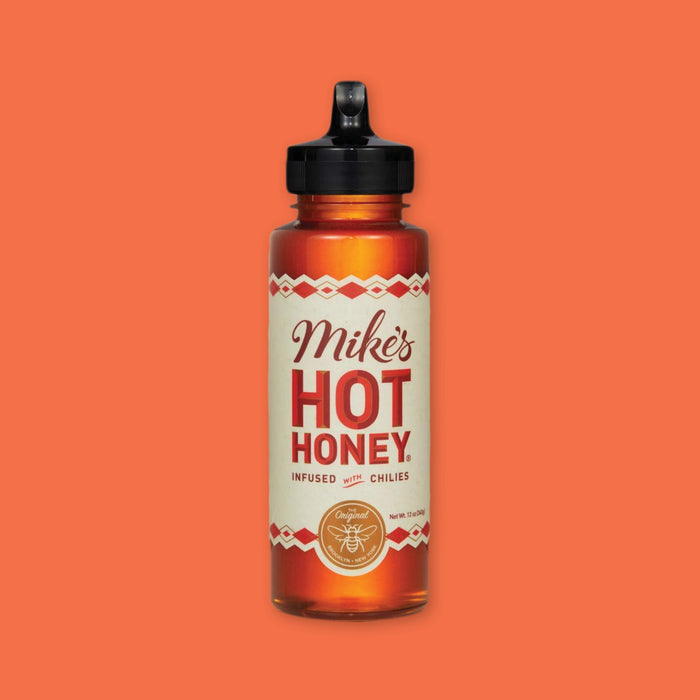 On an orangey-red background sits a bottle. This bottle has a black top and a creamy retro label. It says "Mike's HOT HONEY INFUSED WITH CHILIES" in a maroon and red script and block font. There is a round gold seal at the bottom that says "THE Original BROOKLYN • NEW YORK" with an illustrated outlined bee. Net Wt. 12 oz (340g)