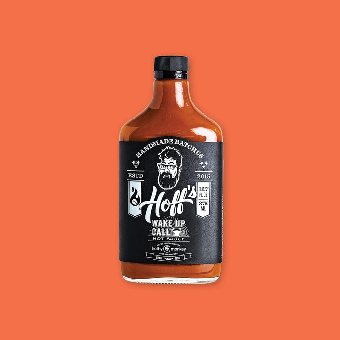 On an orangey-red background sits a bottle. This clear glass bottle has a black top and is filled with orangey-red liquid. On the black label it says "HANDMADE BATCHES" in white, all caps serif font and there is an illustration in white of a man's face with glasses, crazy hair, and a beard. It says "Hoff's WAKE UP CALL HOT SAUCE" in white, script and block font. 12.7 FL OZ 375 ML