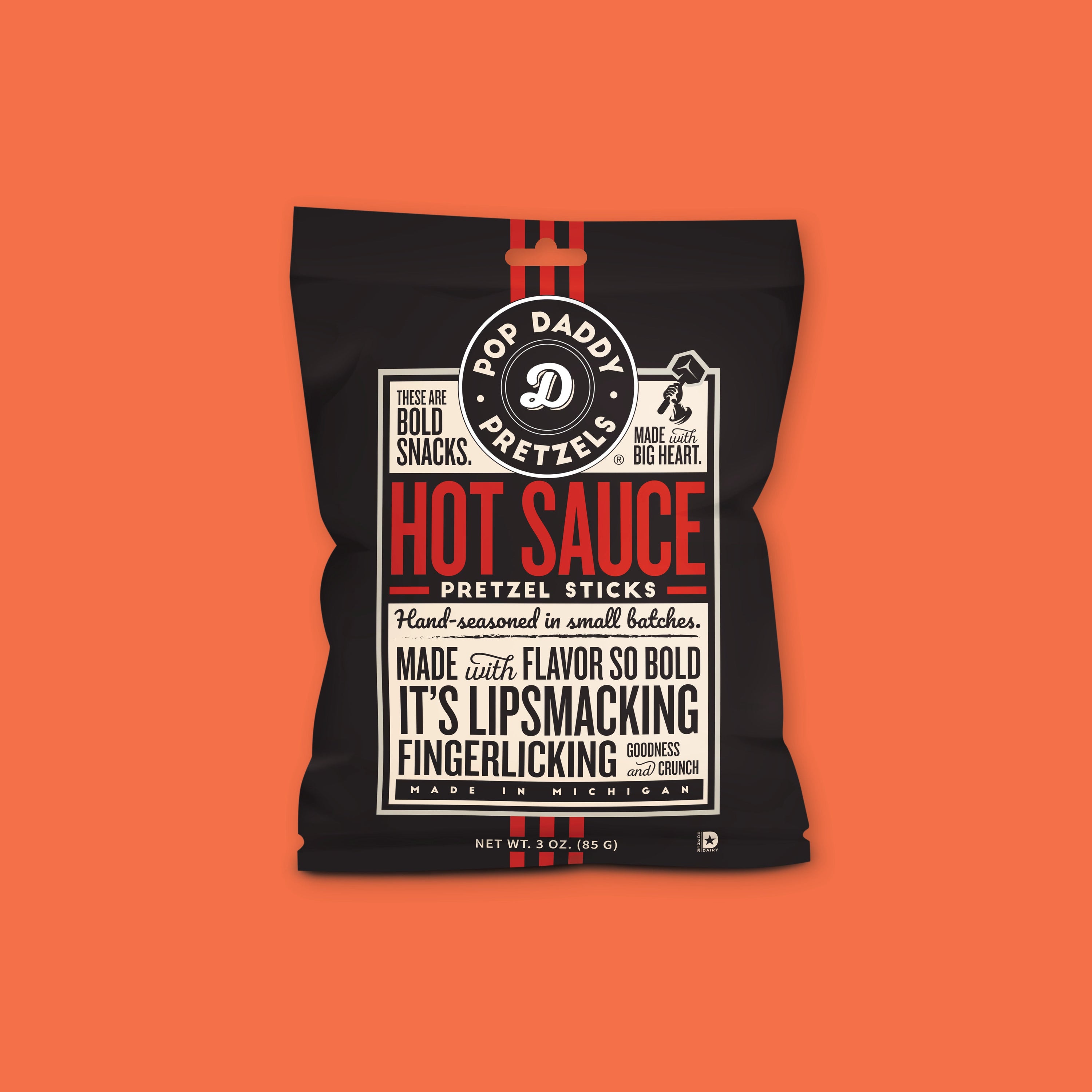 On an orangey-red background sits a package. This package is black with a cream label. There is copy in black and red. It says "POP DADDY PRETZELS," "THESE ARE BOLD SNACKS," "MADE with BIG HEART," "HOT SAUCE PRETZEL STICKS," "Hand-seasoned in small batches," " MADE with FLAVOR SO BOLD IT'S LIPSMACKING FINGERLICKING GOODNESS and CRUNCH," "MADE IN MICHIGAN." NET WT. 3 OZ. (85 G)