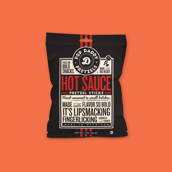On an orangey-red background sits a package. This package is black with a cream label. There is copy in black and red. It says "POP DADDY PRETZELS," "THESE ARE BOLD SNACKS," "MADE with BIG HEART," "HOT SAUCE PRETZEL STICKS," "Hand-seasoned in small batches," " MADE with FLAVOR SO BOLD IT'S LIPSMACKING FINGERLICKING GOODNESS and CRUNCH," "MADE IN MICHIGAN." NET WT. 3 OZ. (85 G)