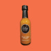 On an orangey-red background sits a bottle. This clear bottle has a black top and is filled with orangey-brown liquid. There is a black circle on the front and in it says "blue collar hot sauce" in white, serif font. Under that says "Black Label Hot Sauce" in white, serif font. NEW WT. 5 FL OZ (150 ML)