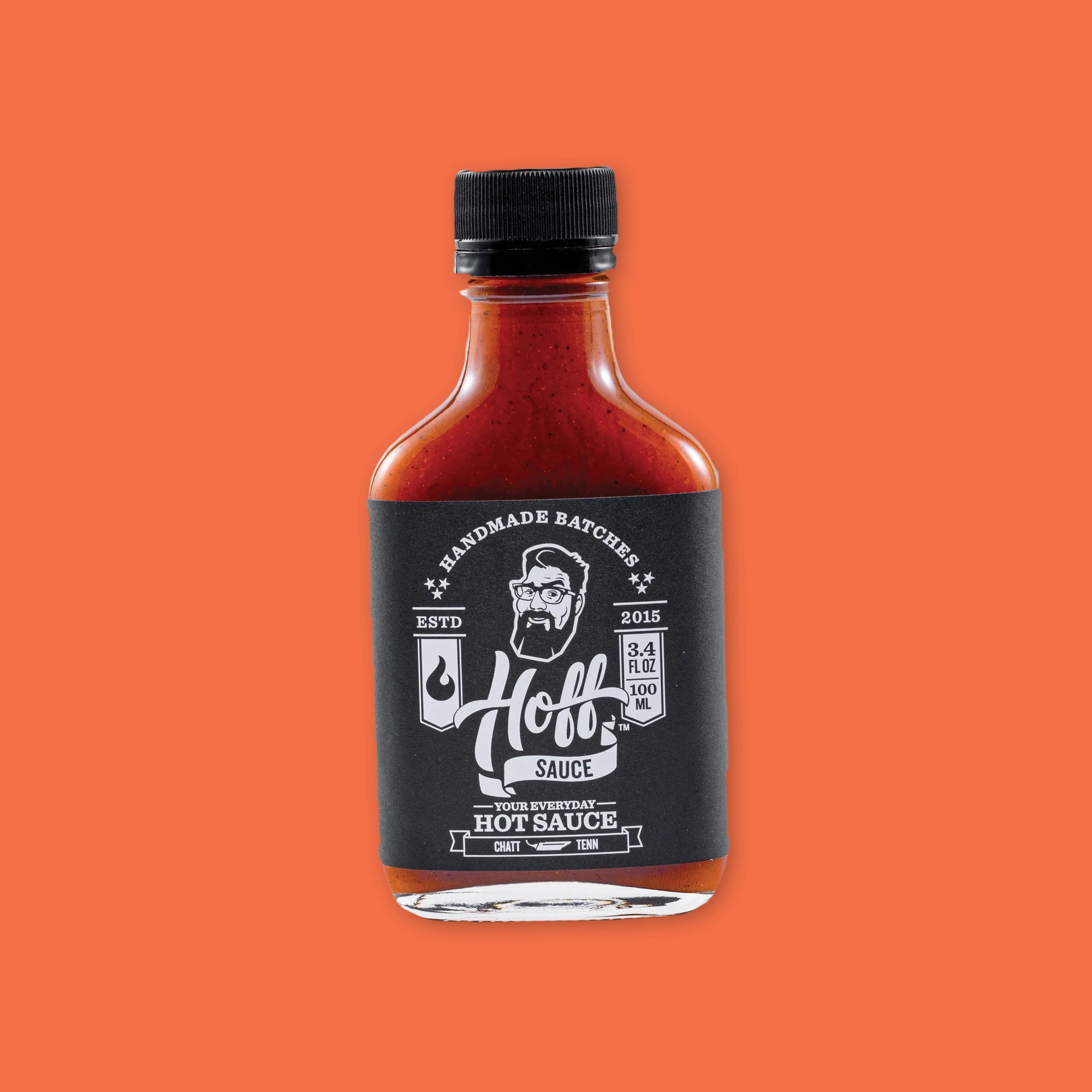 On an orangey-red background sits a bottle. This clear glass bottle has a black top and is filled with orangey-red liquid. On the black label it says "HANDMADE BATCHES" in white, all caps serif font and there is an illustration in white of a man's face with glasses, crazy hair, and a beard. It says "Hoff's SAUCE" in white, script and block font. It also says "YOUR EVERYDAY HOT SAUCE" in white, all caps serif font. 3.4 FL OZ 100 ML