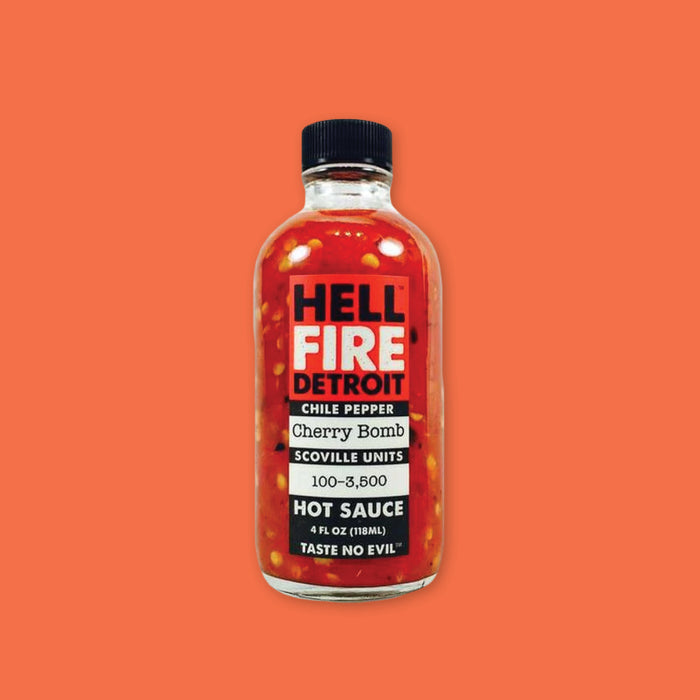 On an orangey-red background sits a bottle. This clear glass bottle has a black top and it is filled with red liquid and pepper seeds. On a red background it says in black and white "HELL FIRE DETROIT" in all caps, block font. It also says on a black background "CHILE PEPPER," "SCOVILLE UNITS," "HOT SAUCE," "TASTE NO EVIL" in white, all caps block font. On a white background it says "Cherry Bomb," "100-3,500" in black, typewriter font. 4 FL OZ (118ML)