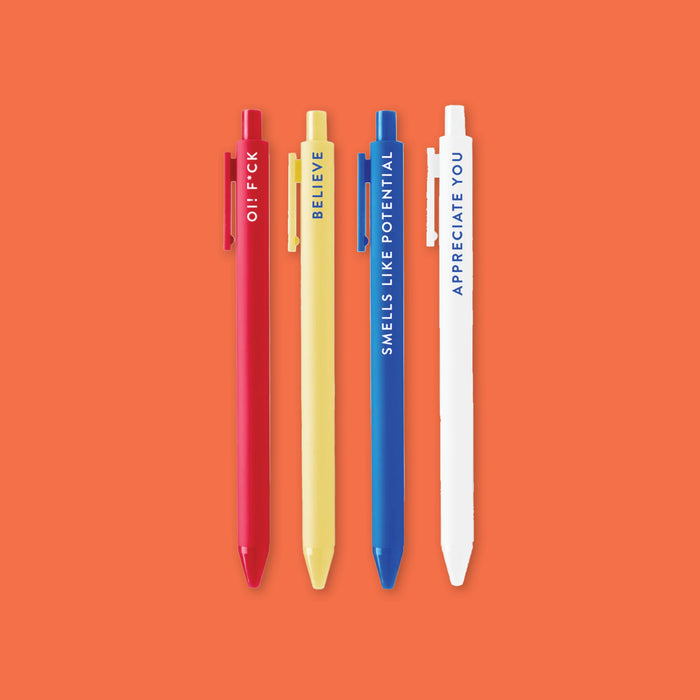 On an orangey-red background sits a wooden tray with an assortment of Ted Lasso themed pens. This photo is a close-up of the set of four retractable gel pens with Ted Lasso sayings. It says "SMELLS LIKE POTENTIAL," "BELIEVE," "APPRECIATE YOU," and "OI! F*CK." They are in blue, light yellow, white, and red.