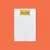 On an orangey-red background sits a notepad. This Ted Lasso inspired white notepad has blue lines on it with an illustration of a yellow "BELIEVE" banner.