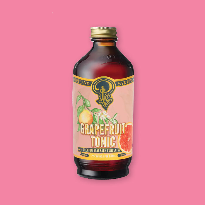 On a bubblegum pink background sits a bottle. This clear glass bottle has a gold lid and a colorful label on the front. It is filled with an amber-red liquid. There is an illustration of grapefruits on a branch with white flowers. It says in gold "PORTLAND SYRUPS" in all caps serif font. It also says "GRAPEFRUIT TONIC" in white and gold, all caps block font. Under that says "PREMIUM BEVERAGE CONCENTRATE" in gold, all caps block font. 12 servings per bottle. 12 Fl oz 355 mL