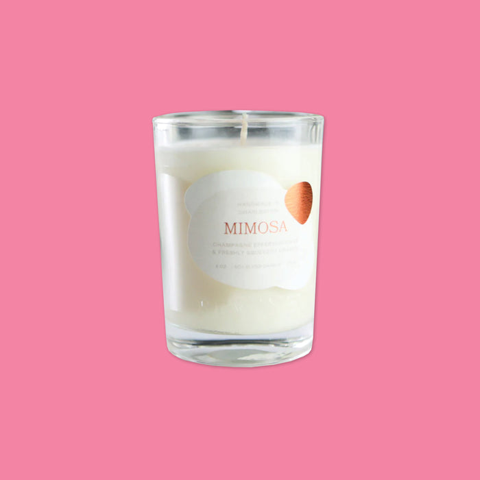 On a bubblegum pink background sits a candle. This clear glass candle has a white label on the front and says "MIMOSA" in orange, all caps serif font.