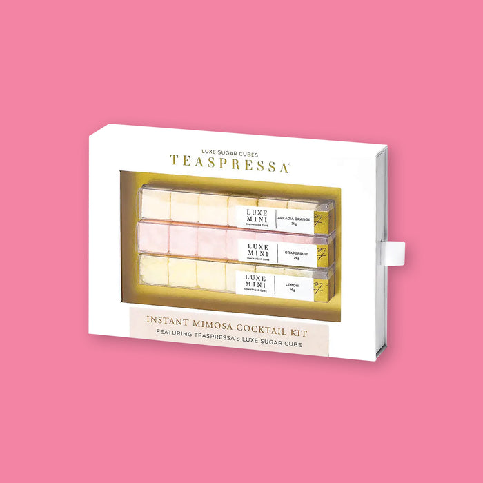 On a bubblegum pink background sits a package. This white package has a clear front and it it are three rows of sugar cubes. The first row is light peach, 'arcadia orange' flavored cubes. The second row is pink, 'grapefruit' flavored cubes. The third row is light yellow, 'lemon' flavored cubes. It says on the top "LUXE SUGAR CUBES TEASPRESSA" in black and gold serif font. On the bottom it says "INSTANT MIMOSA COCKTAIL KIT" in gold, all caps serif font.