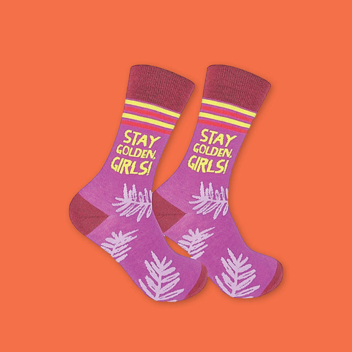 On an orangey-red background sits a pair of purple socks. These Golden Girls inspired socks have dark red, yellow, and red stripes at the top. It has lavender illustrated branches all over the bottom of the socks. It says "STAY GOLDEN, GIRLS!" in yellow, all caps handwritten lettering.