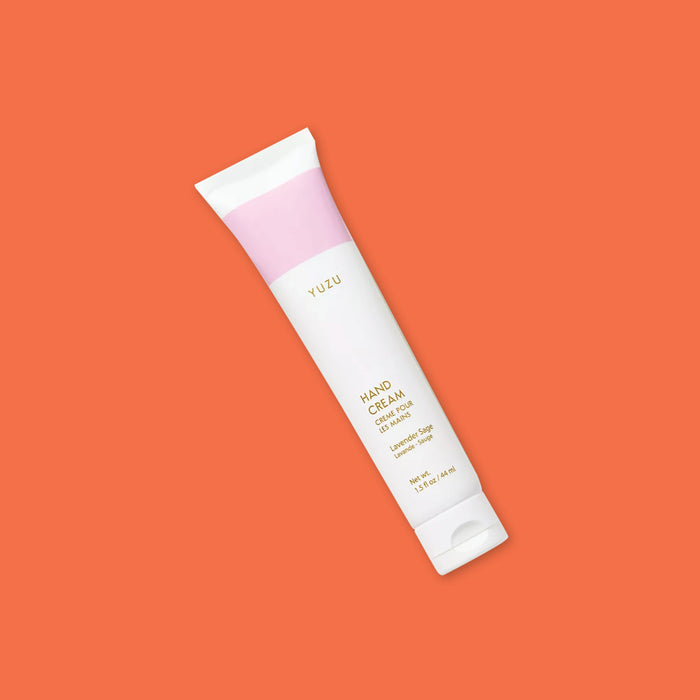 On an orangey-red background sits a white tube, This tube has a bubblegum pink wide stripe at the top. It says "YUZU" and "HAND CREAM CREME POUR LES MAINS" in gold, all caps font. It is a 'Lavender Sage' scent. Net wt. 1.5 fl oz / 44 ml