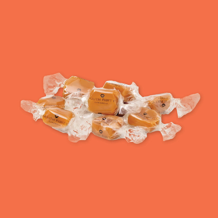 On an orange backgorund sits a pile Gourmet Caramels each wrapped in clear cello with "Mouth Party Pumpkin Spice" on each wrapper.