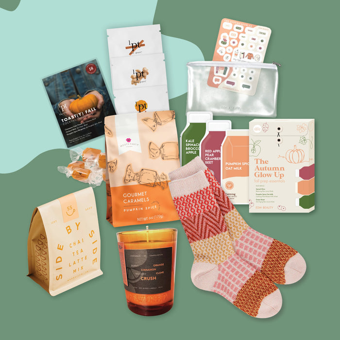 On a green and aqua background jauntily sits an assortment of fall products for a pumpkin spice themed gift box, including a cozy scarf, caramels, face masks and more.