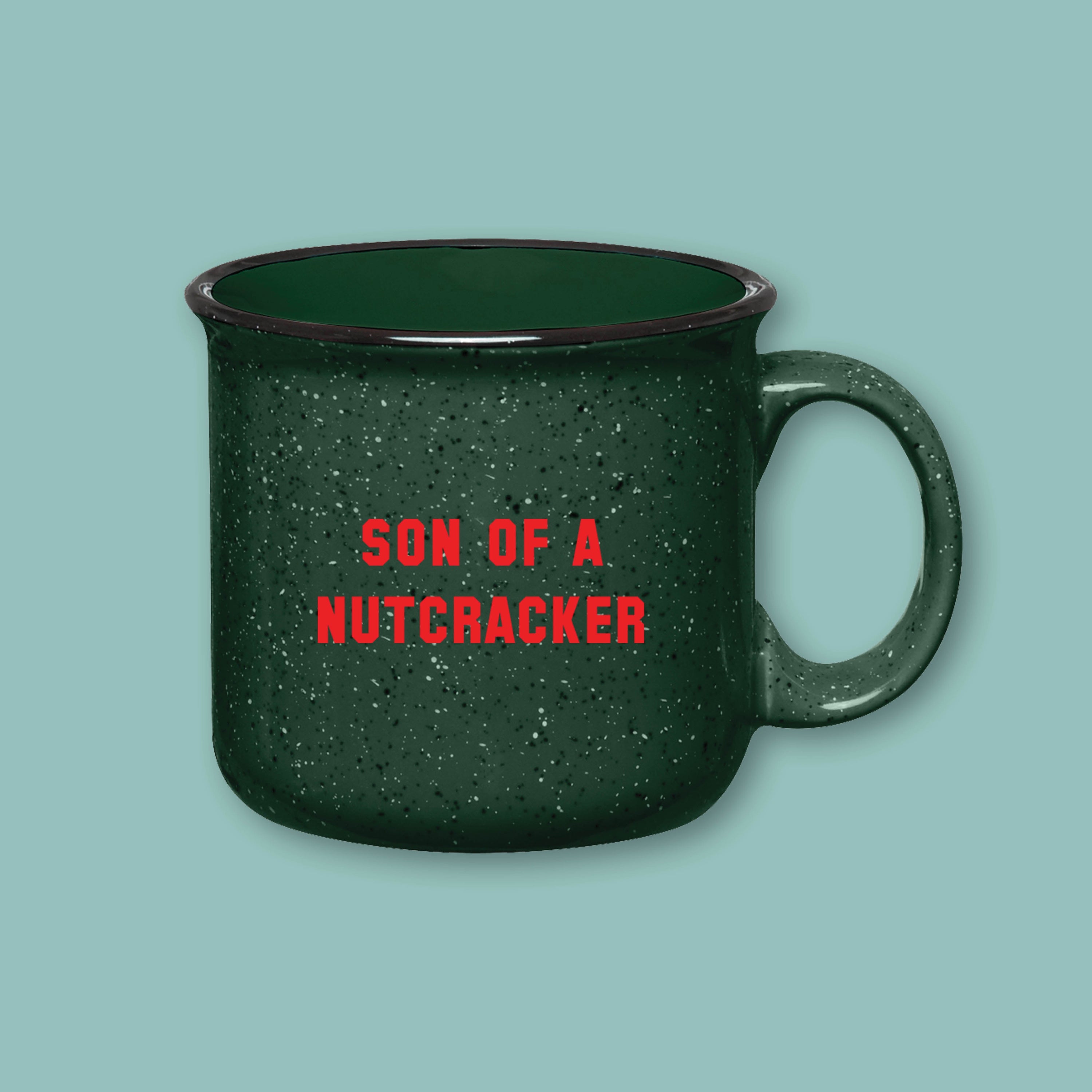 On a sage green background sits a campfire mug. This Elf inspired mug is dark green with black and white specks and it's say in red lettering "SON OF A NUTCRACKER"