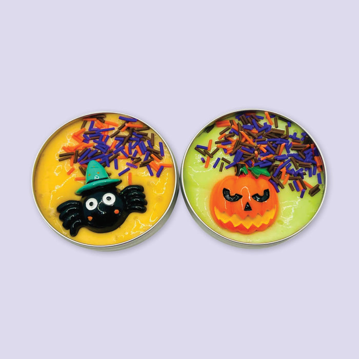On a lavender background sits two small tins of Halloween slime -- one yellow with a witchy spider charm and sprinkles, the other lime green with an angry orange pumpkin charm and sprinkles.